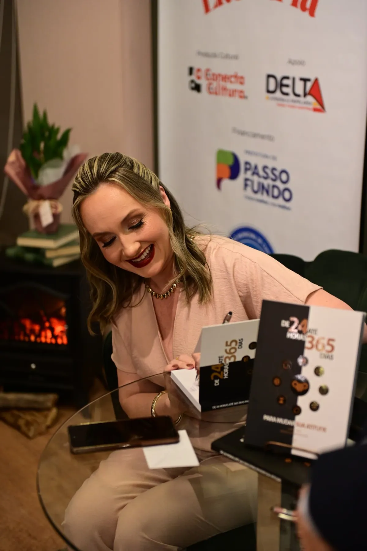 Graciela Oliveira Roth Colle sorrindo enquanto autografa o livro, com exemplares da 3ª edição sobre a mesa